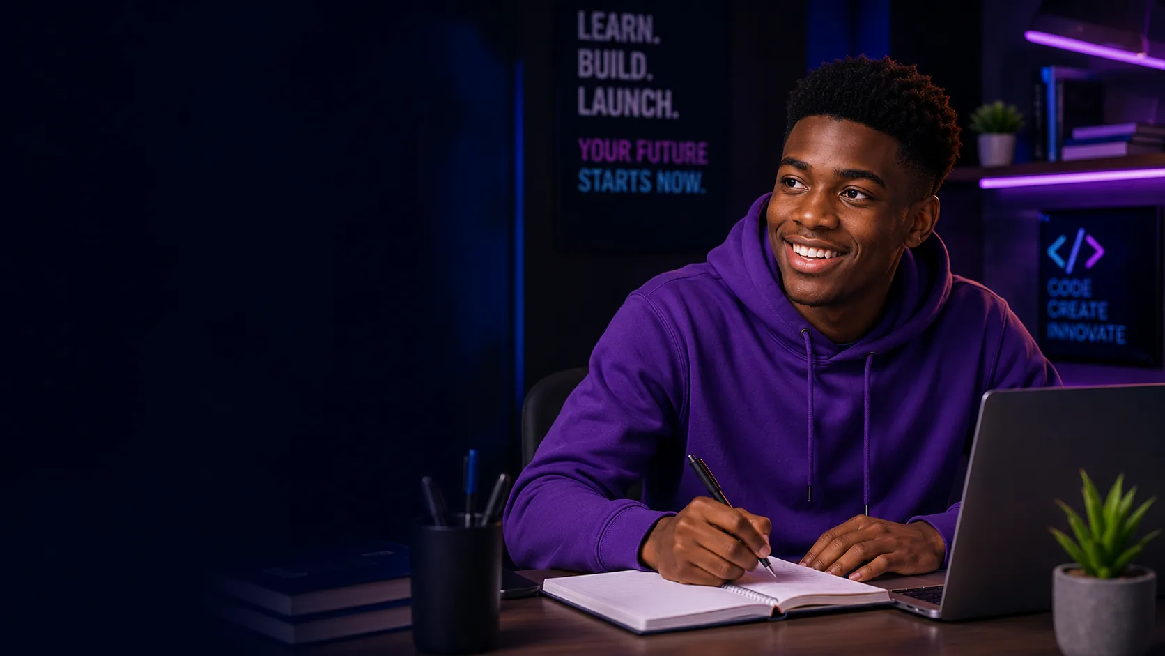A smiling young man in a purple hoodie writing in a notebook beside a laptop